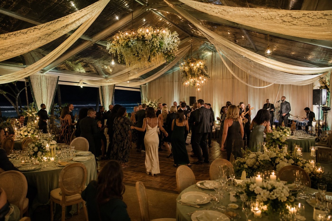 Guests dancing beside a live band inside a fabric-lined glass wedding tent